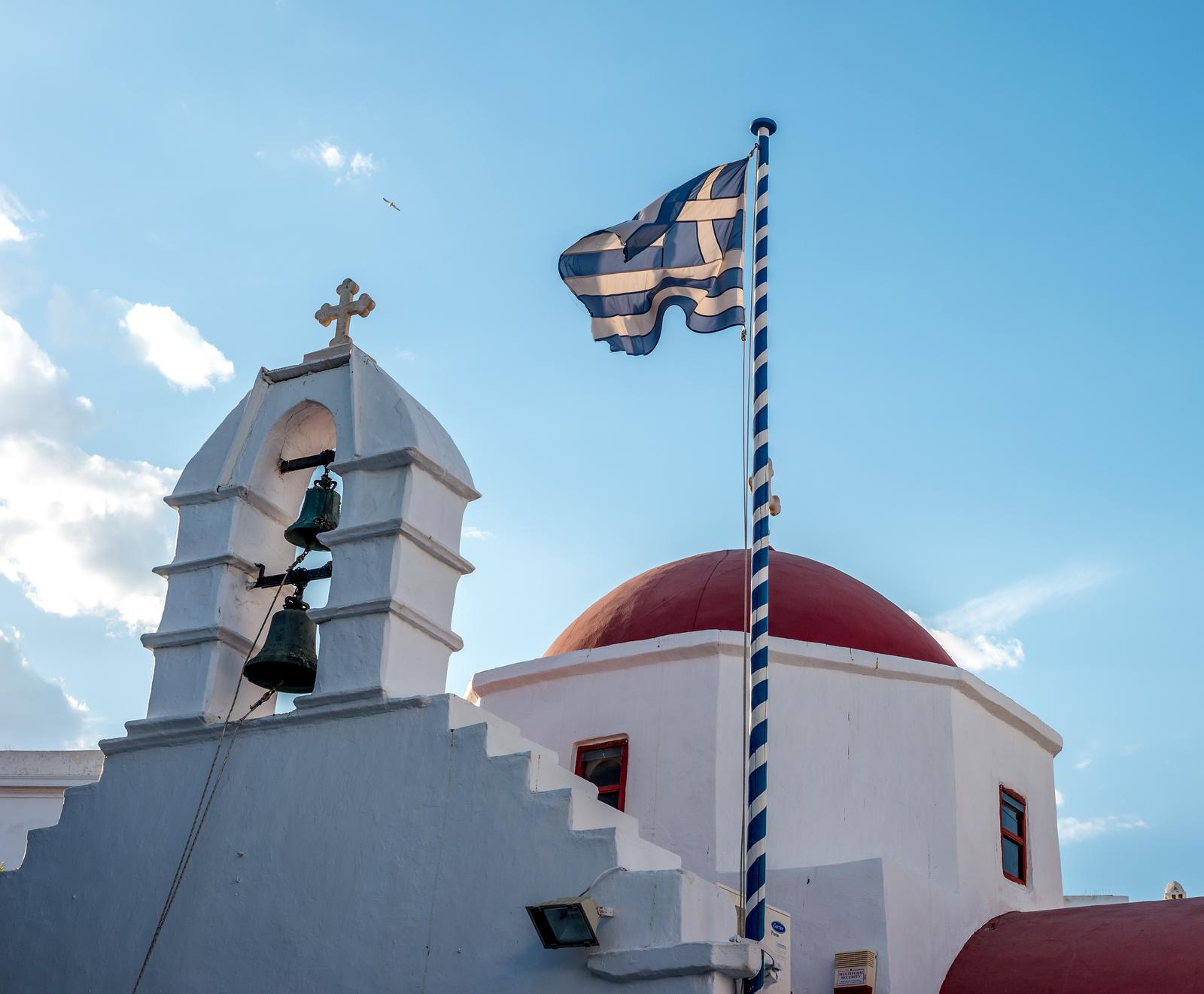 Agia Kyriaki Orthodox church with red dome and Greek flag waving against blue sky on the Mykonos island, Greece